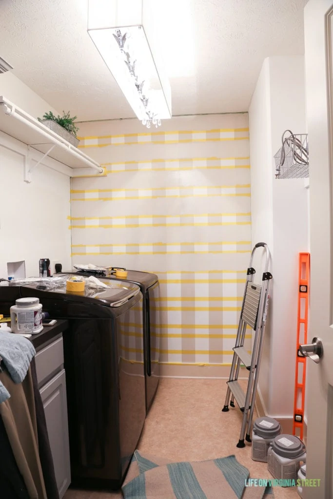 The buffalo checked wall all taped up in laundry room with stepladder against wall.