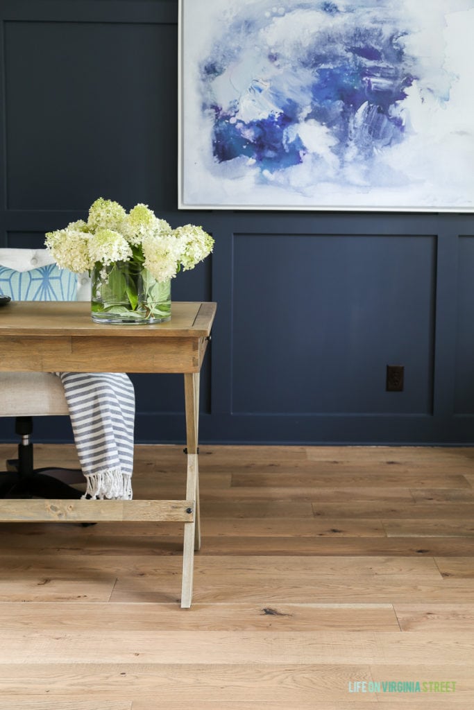 Office with natural wood desk and navy blue board and batten walls.  There is a clear vase on the desk with hydrangeas in it.