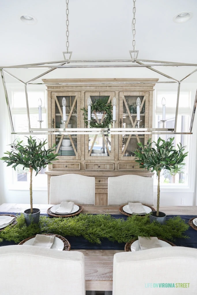 Fall dining room with blue and white striped rug, natural wood table, linen chairs, linen roman shades, reclaimed wood china cabinet, olive leaf topiaries and beach art. Light fixture is the Visual Comfort Darlana Linear Pendant.