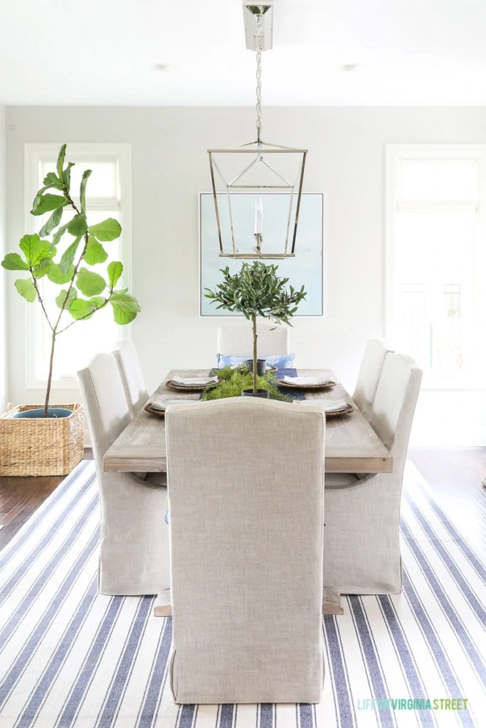 Neutral dining room with blue and white striped rug, natural wood table, linen chairs, fiddle leaf fig tree, olive leaf topiaries and beach art. Light fixture is the Visual Comfort Darlana Linear Pendant.