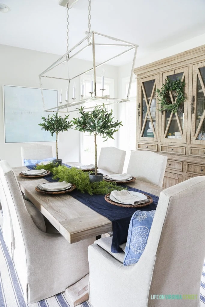 Fall dining room with blue and white striped rug, natural wood table, linen chairs, reclaimed wood hutch with olive wreath, olive leaf topiaries and beach art. Light fixture is the Visual Comfort Darlana Linear Pendant.