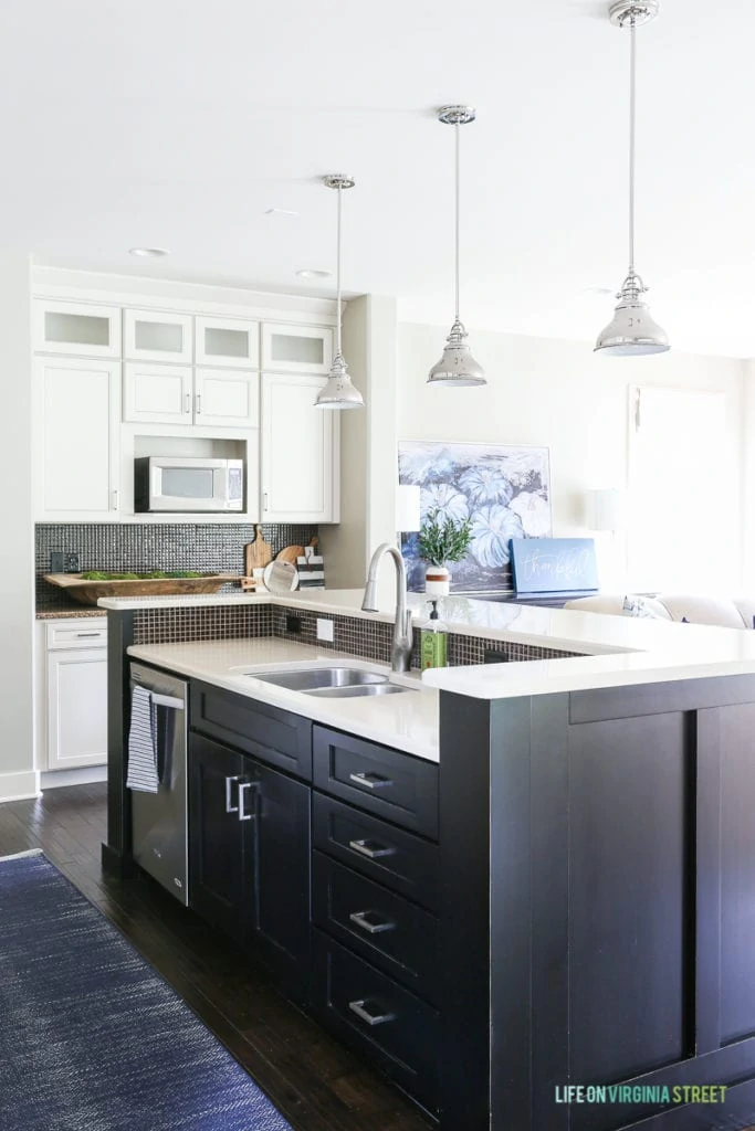 White kitchen with black island, navy blue herringbone rug, and chrome pendants over island.