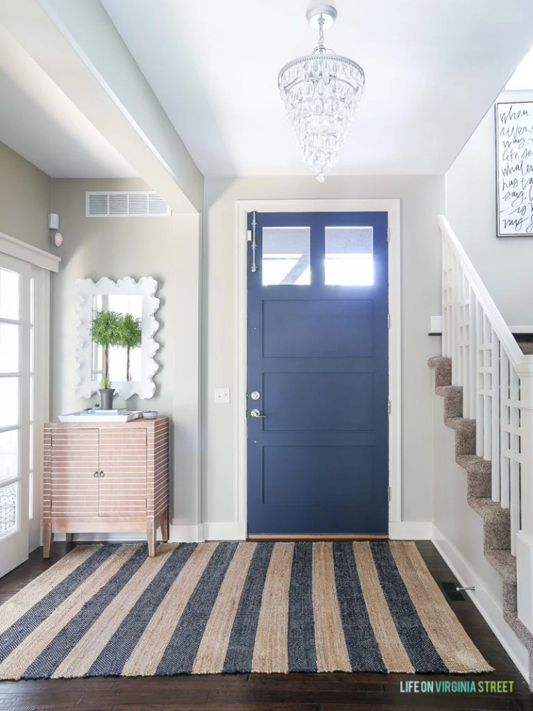 Entryway with navy blue front door, jute and navy striped rug, mossy topiary, white coral mirror, crystal chandelier and striped wood cabinet. Wall color is Behr Castle Path and door color is Benjamin Moore Hale Navy.