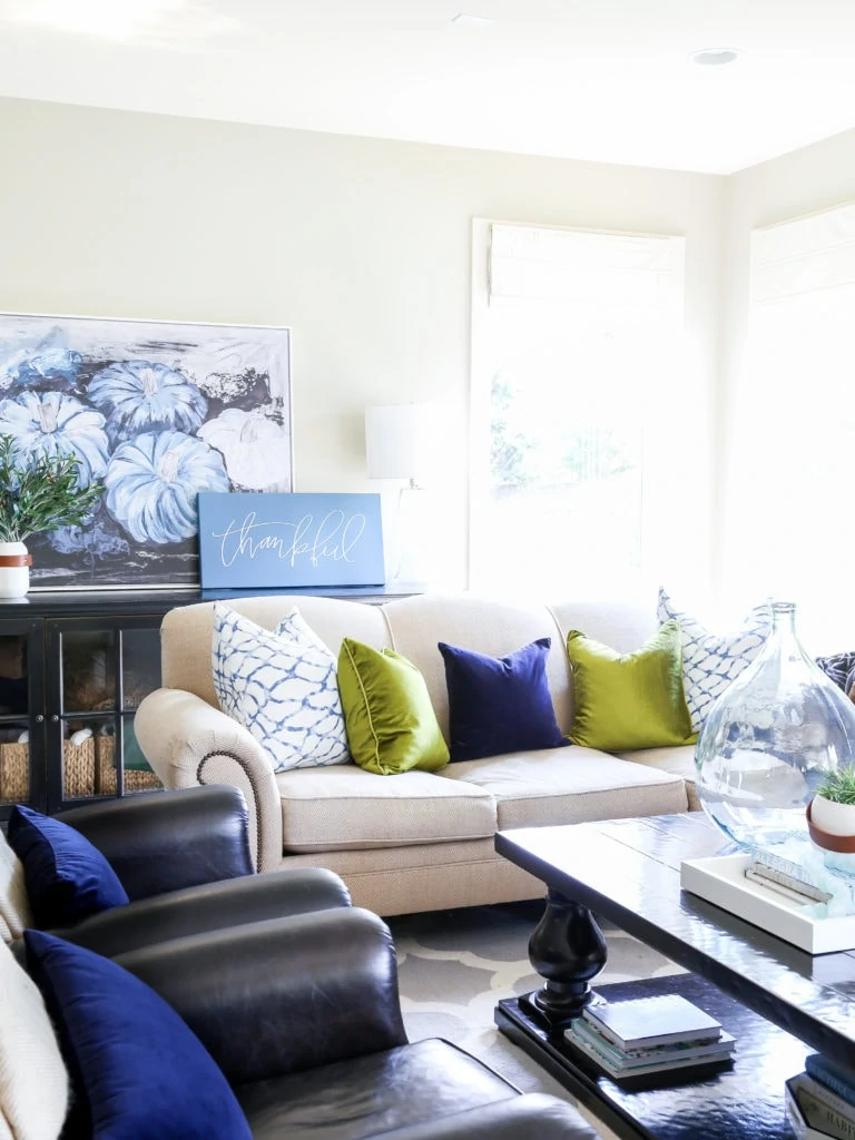 Fall living room with blue pumpkin artwork, neutral sofa, chartreuse and navy blue velvet pillows, olive leaf branches and wooden coffee table.