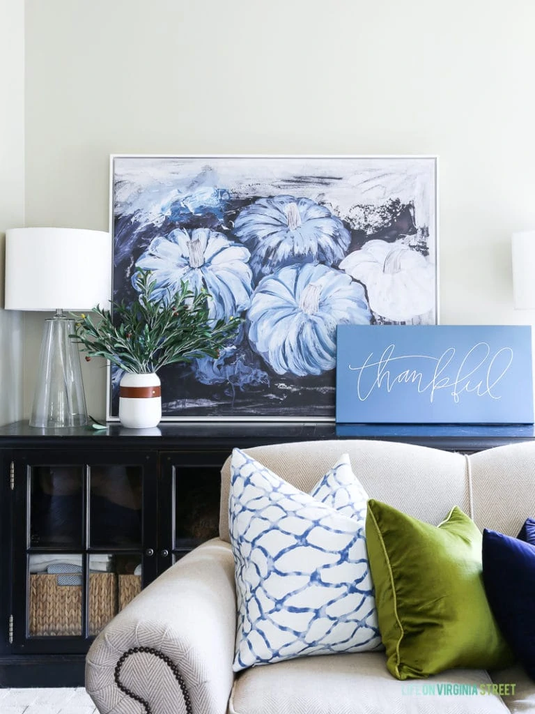 Fall living room with blue pumpkin artwork, neutral sofa, chartreuse and navy blue velvet pillows, glass lamps, and olive leaf branches.