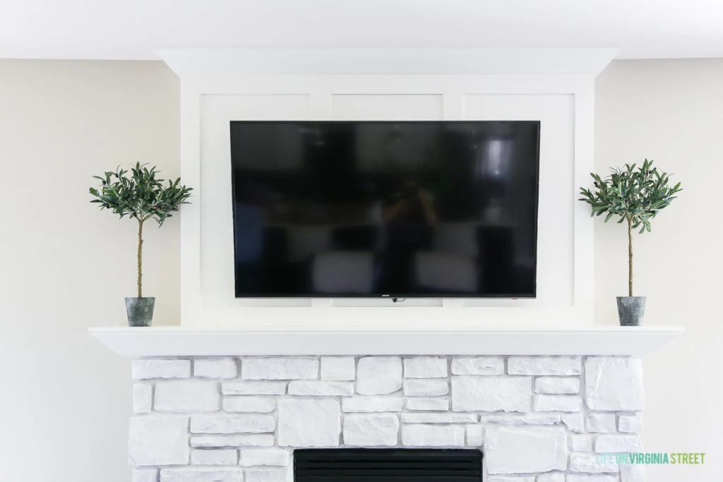 White stone fireplace with topiaries on the mantel and a TV hanging above it.