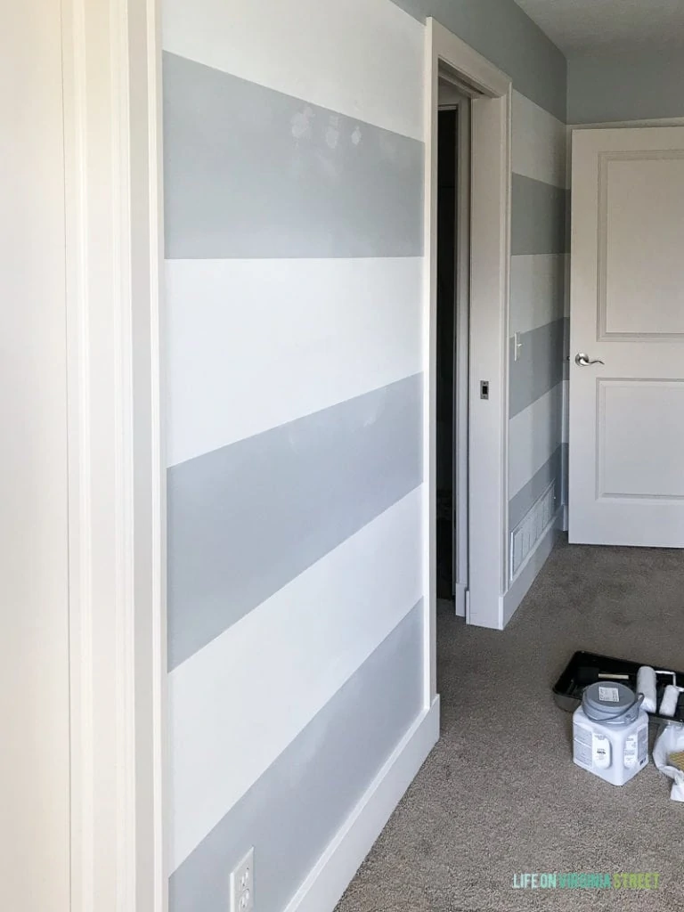 Blue and white striped wall in bedroom with paint can and roller on floor.