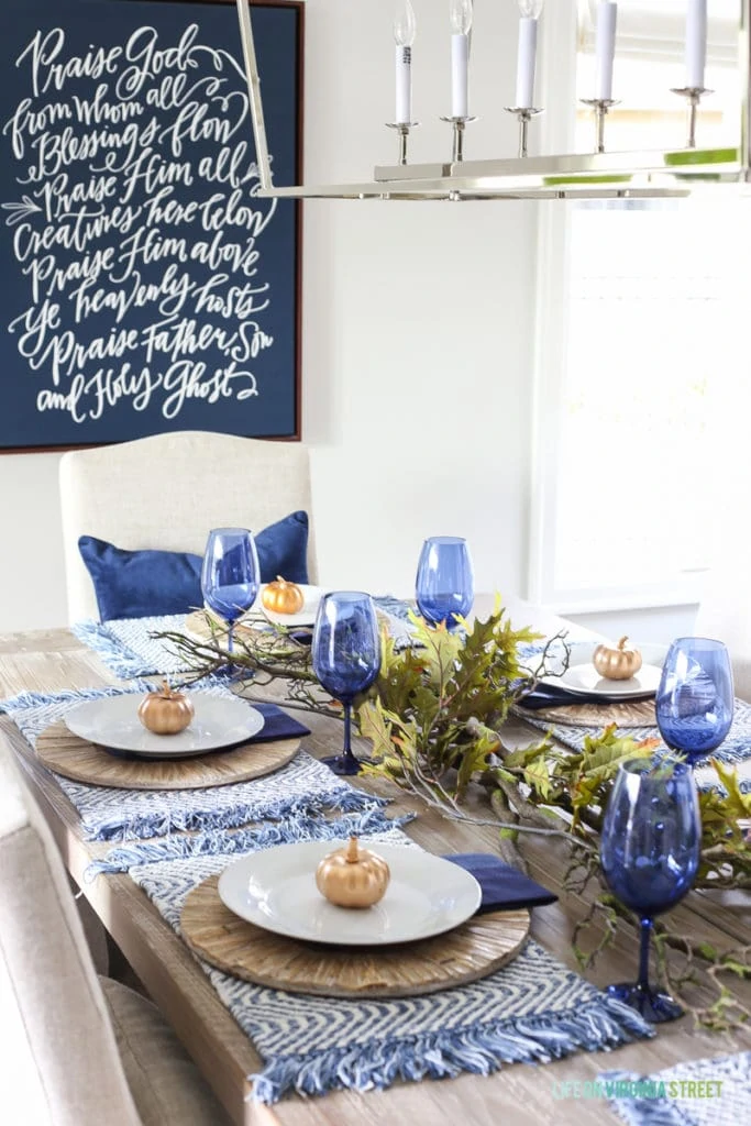 Blue and white Thanksgiving dining room with navy blue doxology canvas, Darlana linear pendant, blue wine glasses, navy blue velvet pillow, oak leaves and branches centerpiece, reclaimed wood table, and linen chairs.