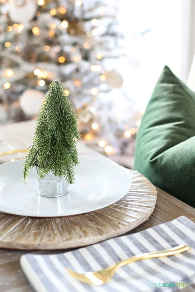 Woodland Christmas dining room with a navy blue striped rug, linen chairs, reclaimed wood dining table, 'O Come Let Us Adore Him' canvas art, and a flocked Christmas tree.