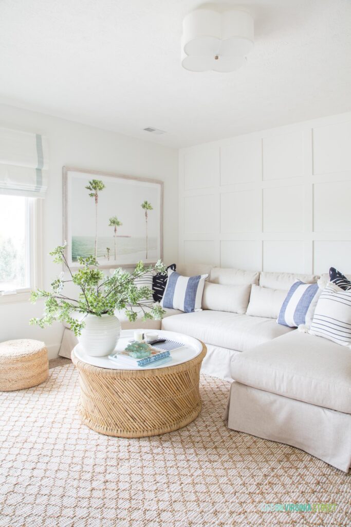 A living room with a round coastal coffee table, linen sectional, jute rug, palm tree art, board and batten grid wall, and blue and white decor.