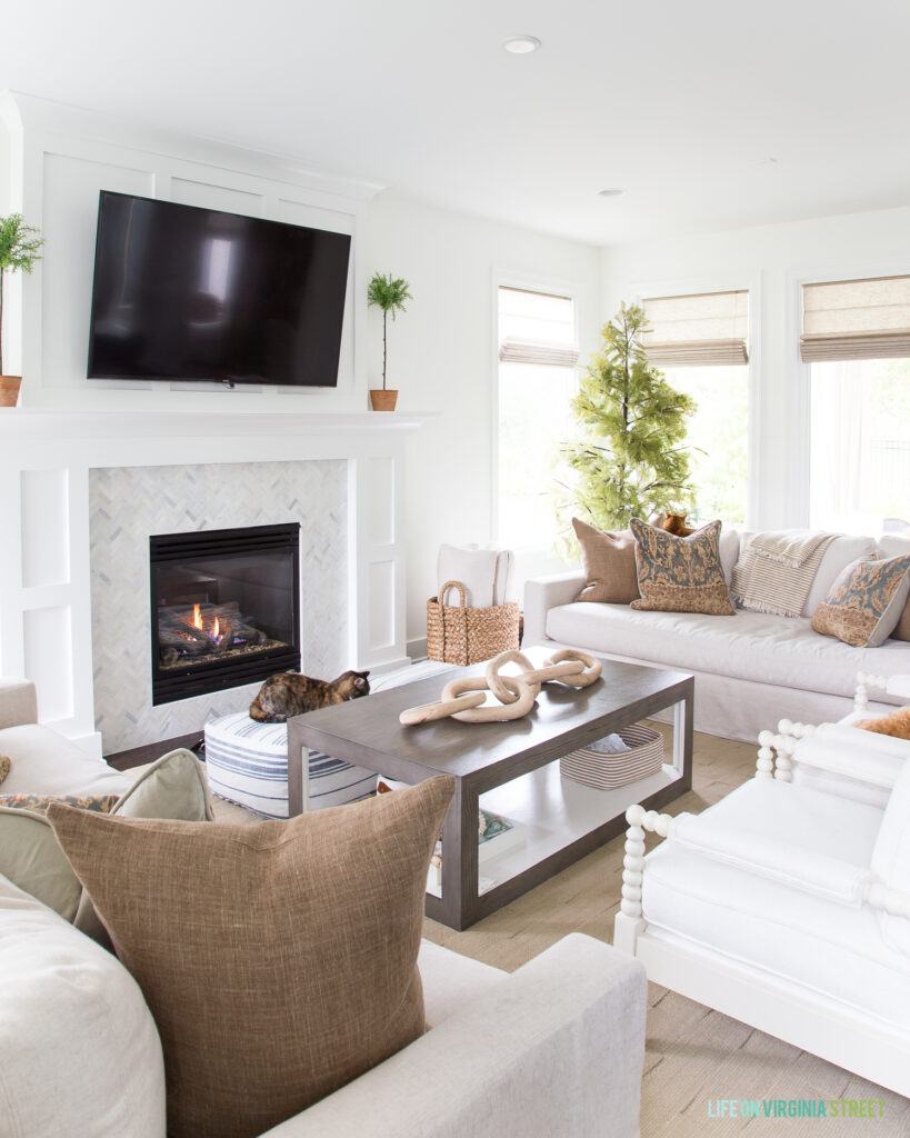 A modern coastal coffee table in a living room with white walls, marble fireplace surround, white spindle chairs, and brown linen pillows.