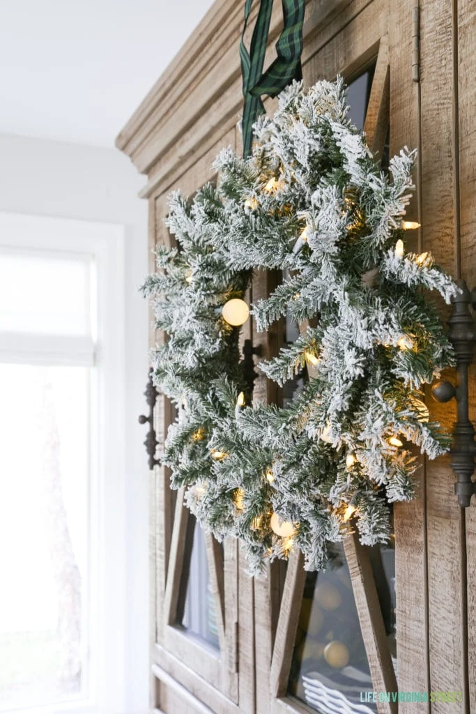 A flocked wreath hanging with plaid ribbon on a reclaimed wood hutch.