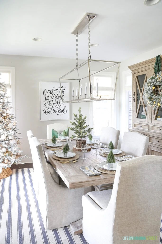 Coastal dining room with blue and white striped rug, linen chairs, reclaimed wood dining table, Darlana linear pendant and a flocked Christmas tree.