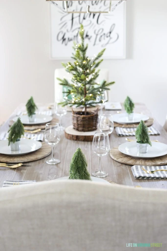 A coastal woodland tablescape with a reclaimed wood table, linen chairs, green velvet pillow, and Christmas trees.