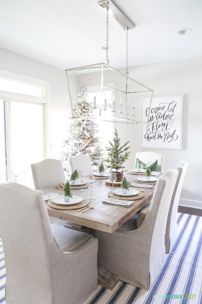 Woodland Christmas dining room with a navy blue striped rug, linen chairs, reclaimed wood dining table, 'O Come Let Us Adore Him' canvas art, and a flocked Christmas tree.
