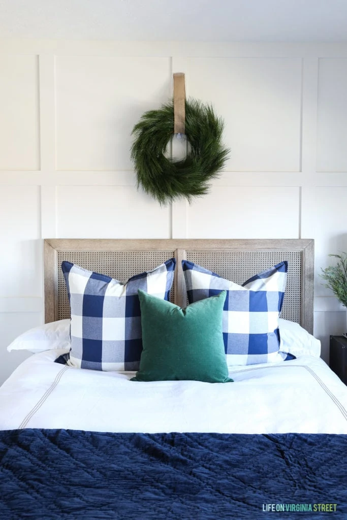 Coastal style bedroom with whitewashed cane wood bed, white bedding, wreath over bed, navy blue velvet coverlet, dark green velvet pillow and buffalo check pillows. Simple Christmas decorating ideas.