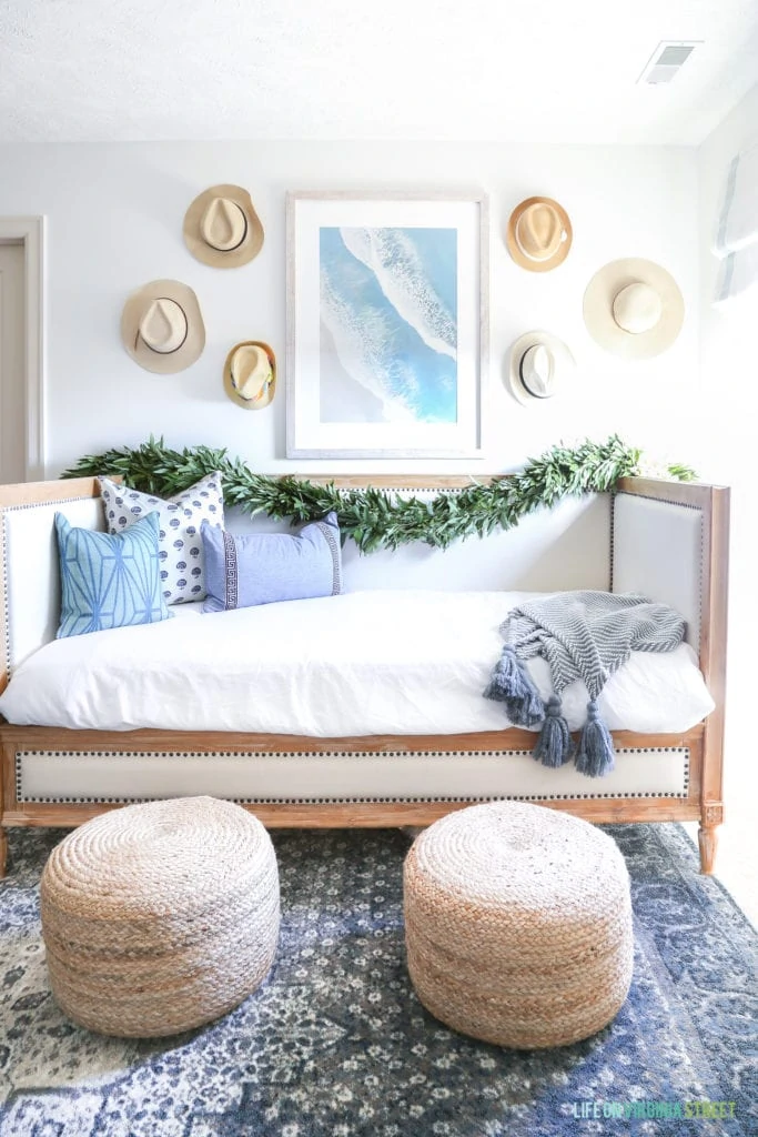 Wood and linen daybed, blue and brown vintage style rug, jute poufs, beach art, and hats on the wall.