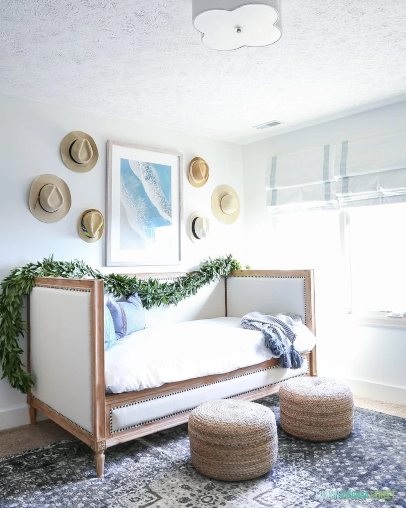 Daybed with white bedding, jute poufs, navy blue vintage rug, scalloped light fixture, beachy art and hats on the wall.