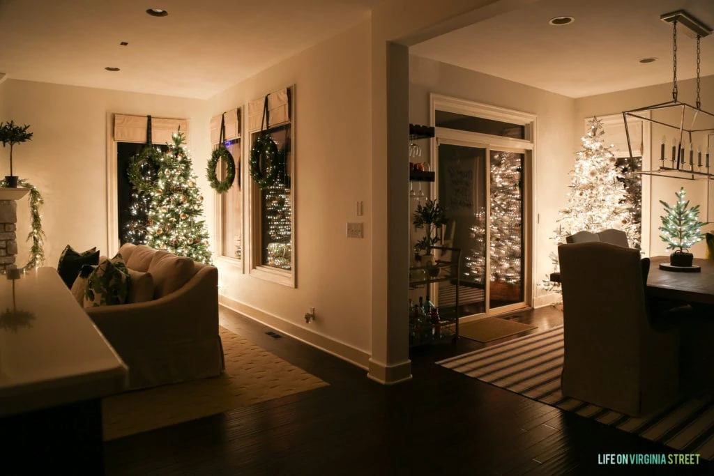 A split view of the living room and the dining room both have Christmas trees in the corner, one is green and the other is white.