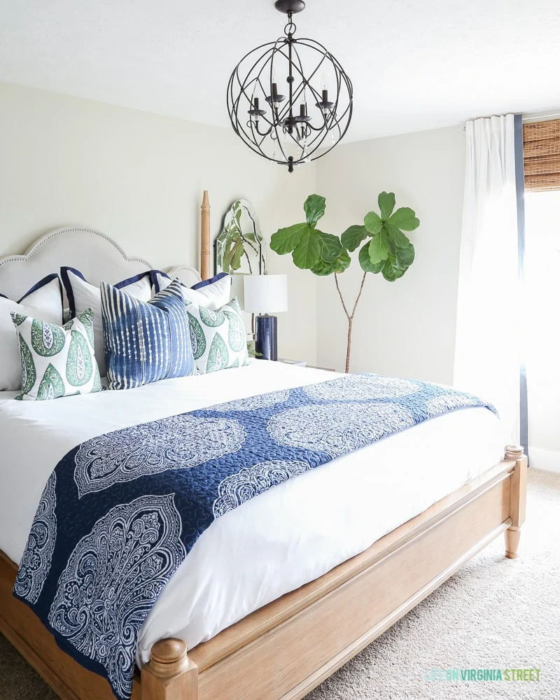 Bedroom with light gray walls, blue and white bedding, orb chandelier, natural woven shades and a fiddle leaf fig tree.