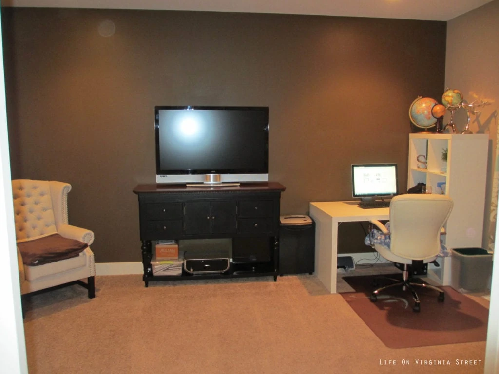 The office with dark brown walls, a small desk and armchair.  A dark room.