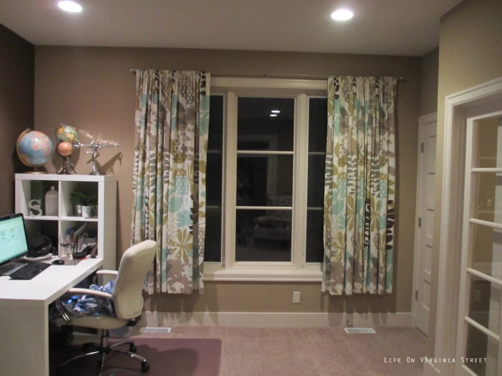 The office with a small white desk and floral curtains and beige walls.