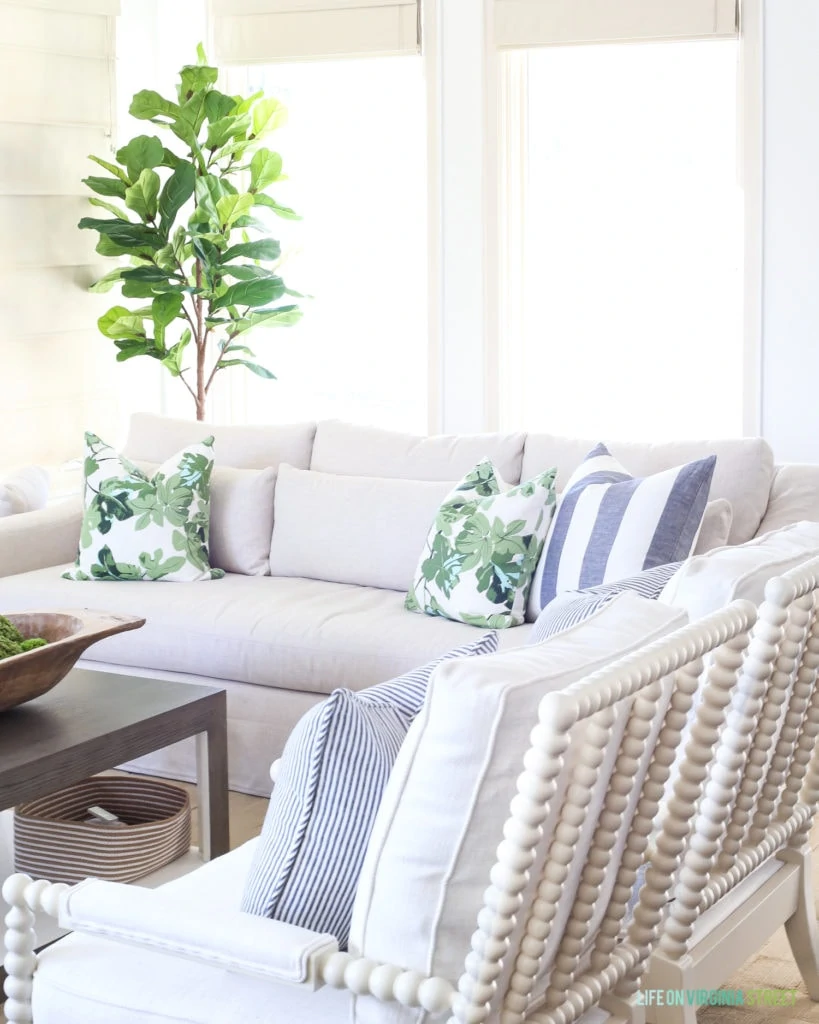Living room with neutral linen sofa and white spindle chairs, fig leaf pillows, navy striped pillows and a faux fiddle leaf fig tree. Wall color is Benjamin Moore Simply White.