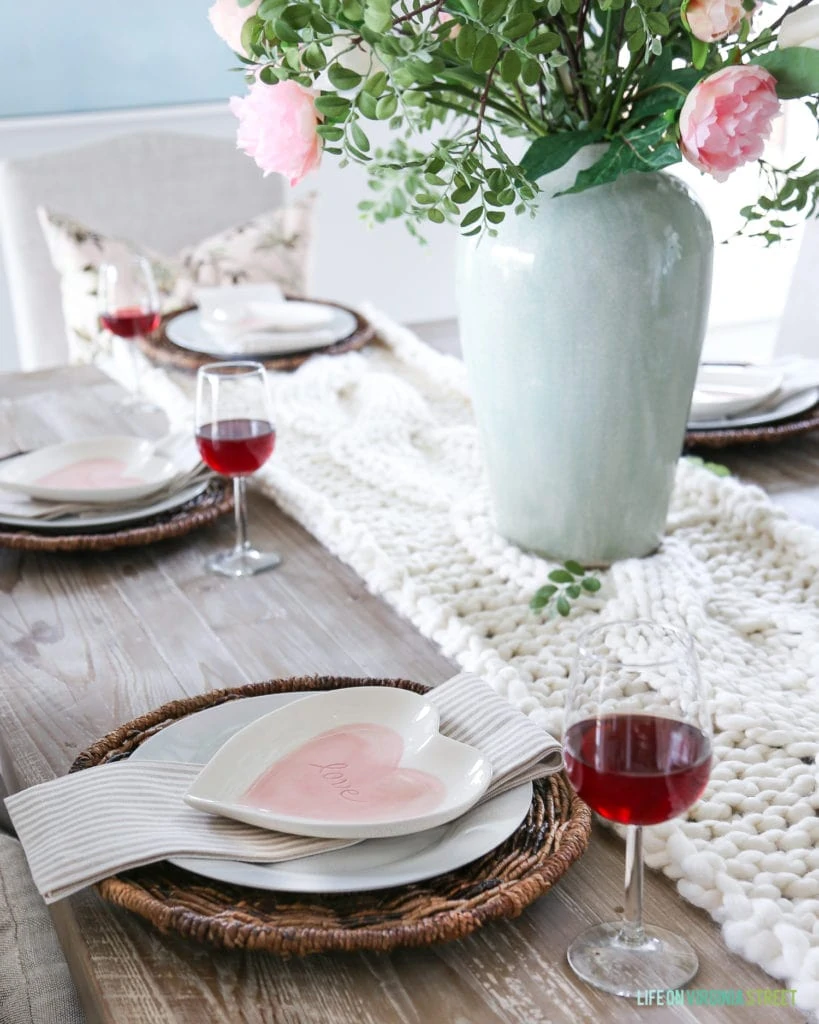 A Valentine's Day place setting on a reclaimed wood table, with celadon green vase, chunky knit table runner, and pink heart shaped plates.