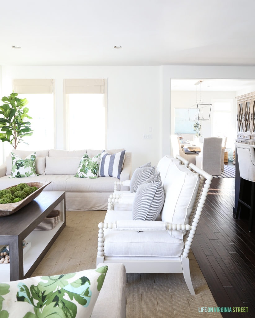 White living room with white spindle chairs, oatmeal linen sofas, striped pillows, and fig leaf pillow. Walls are painted Benjamin Moore Simply White.