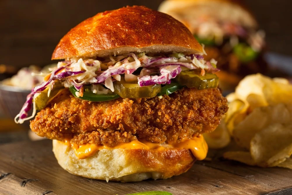 A chicken burger with pickles, onions and sauce on a cutting board.