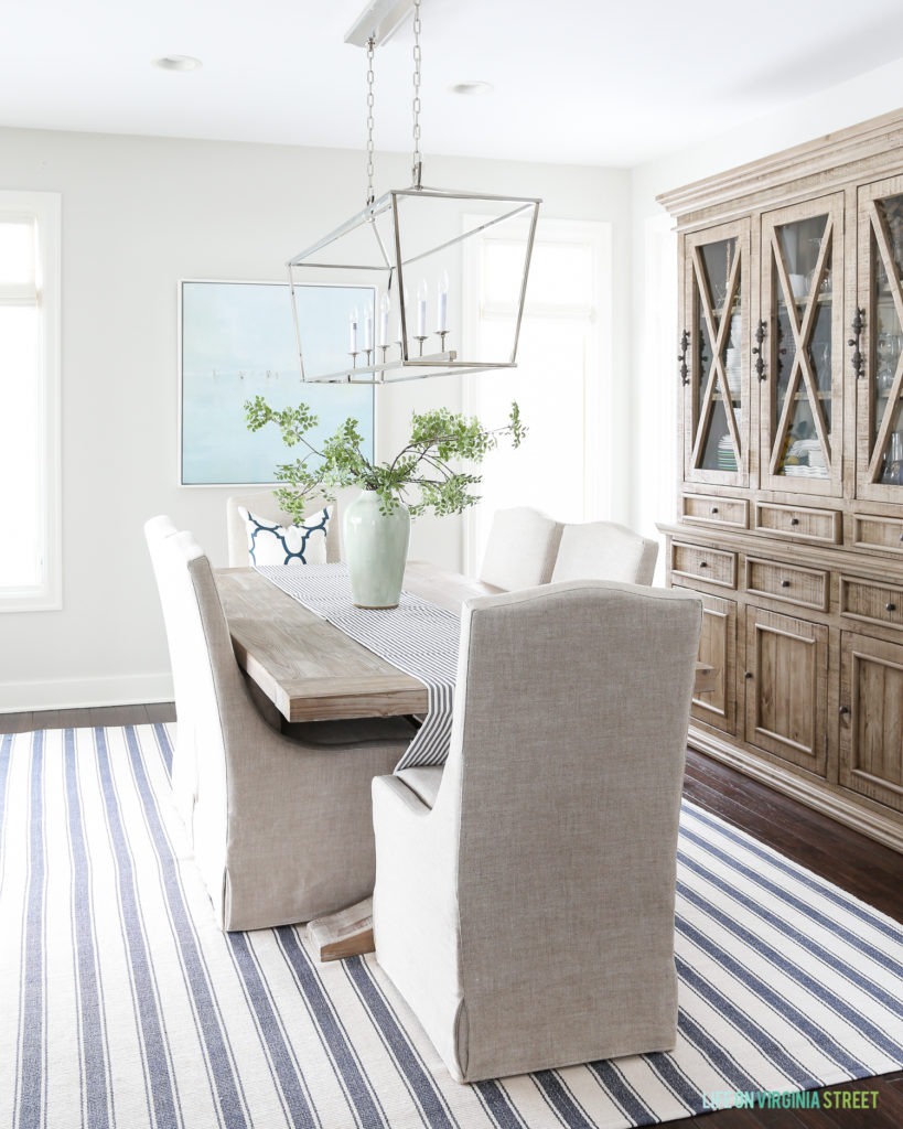 Visual Comfort Darlana Linear Pendant Light hanging in the dining room with a wood table, fabric chairs, and blue and white striped rug.