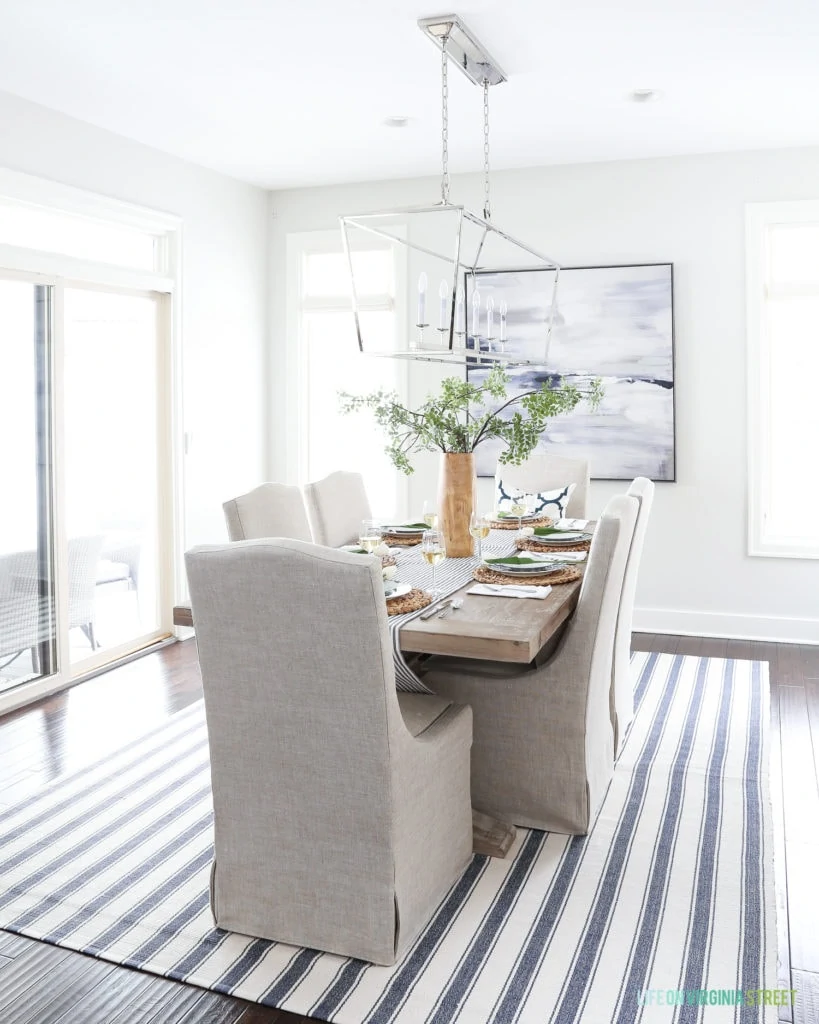 Dining room with Behr Silver Drop paint and navy blue and white accents. Linen chairs, reclaimed wood dining room table, navy blue striped rug, and Darlana linear pendant light fixture.