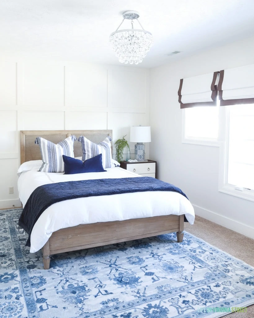 A bedroom with a driftwood rattan headboard, blue and white striped pillow, a crystal chandelier, ribbon trimmed roman shades, and a blue patterned rug.