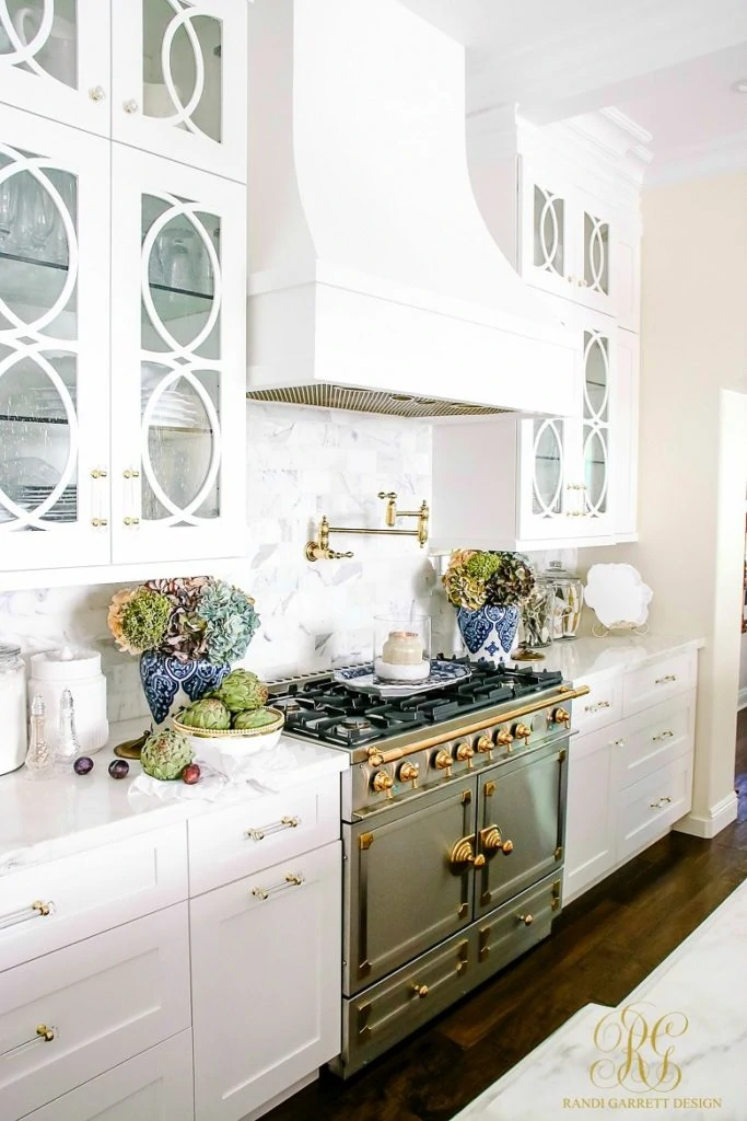 A white kitchen with a stove with gold details and a gold pot filler.   There is flowers and fruit on the counter.