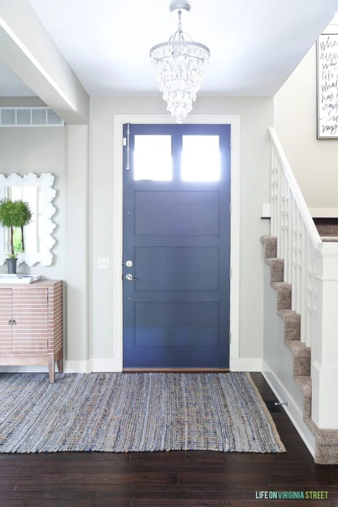 Spring entryway with Benjamin Moore Hale Navy front door. Walls are Behr Castle Path. A small rug in front of the front door and a chandelier in the hallway.