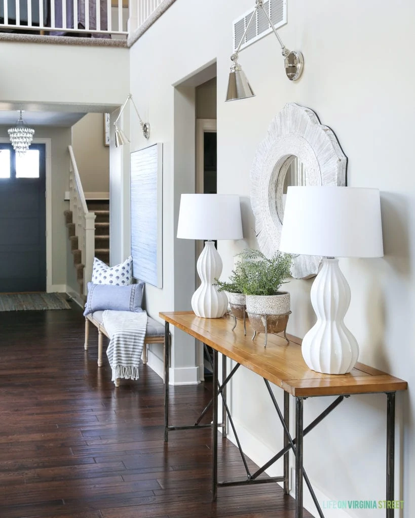 Spring entryway with white orb lamps, wood and iron console table, maidenhair ferns, chrome swing arm sconces, linen and wood bench and a navy blue front door. 