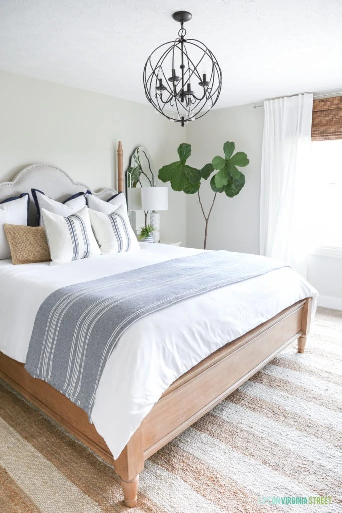 A coastal farmhouse style bedroom with a striped rug, orb chandelier, navy blue and white striped blanket, grainsack pillows and fiddle leaf fig tree.