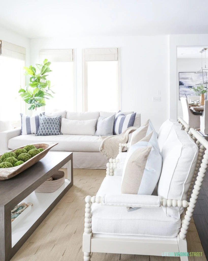 Bright coast living room with linen slipcovered sofas, blue and white accents pillows, white spindle chairs, and a modern coffee table with a large dougbowl for decoration.