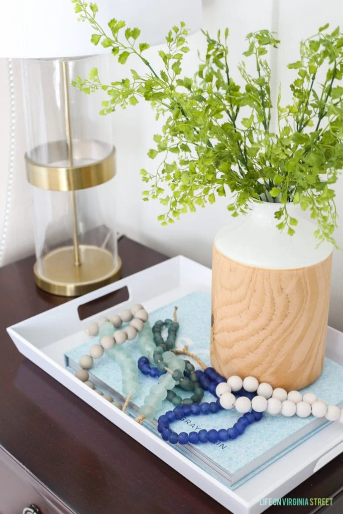 A tray with beads on a string in it plus a vase with greenery in it beside the bed.