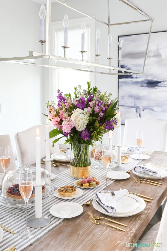 A dining room table with purple, white and pink floral arrangement, white dishes and gold cutlery.