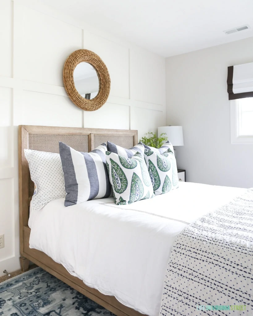 Coastal style bedroom with white linens, striped pillows, paisley accents, a woven rope mirror, board and batten wall and blue patterned rug.