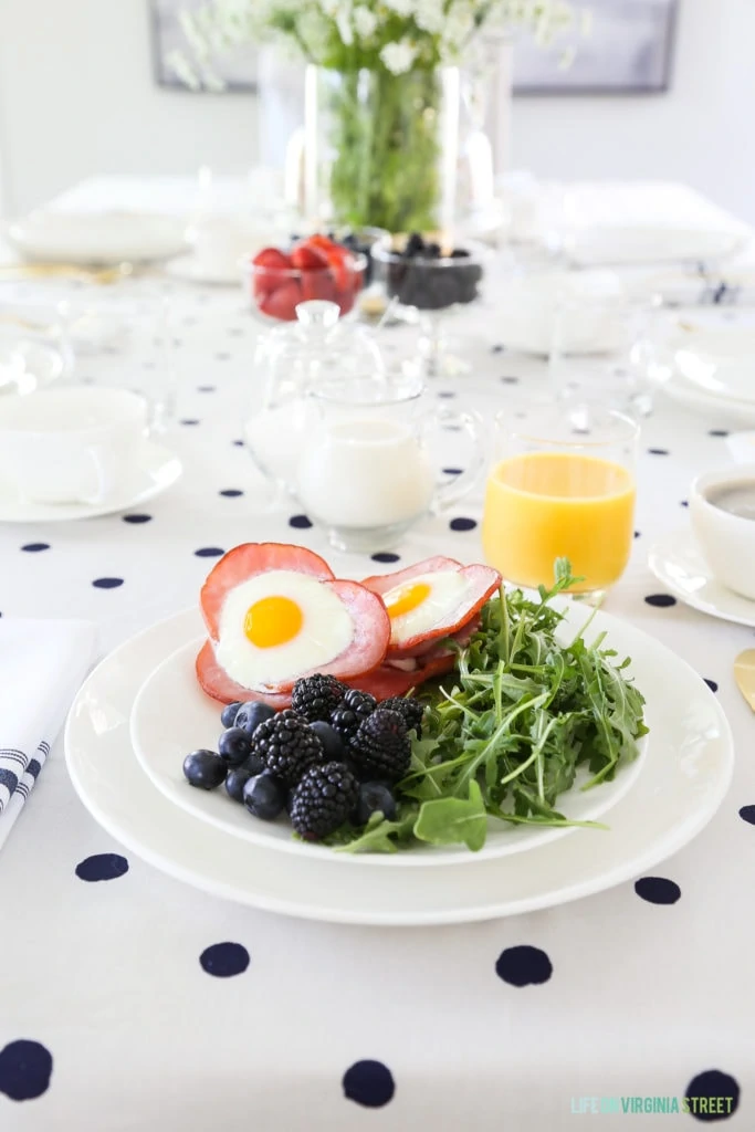 The plate of food with the eggs, bacon, berries and greens and a glass of orange juice on a polka dotted tablecloth.