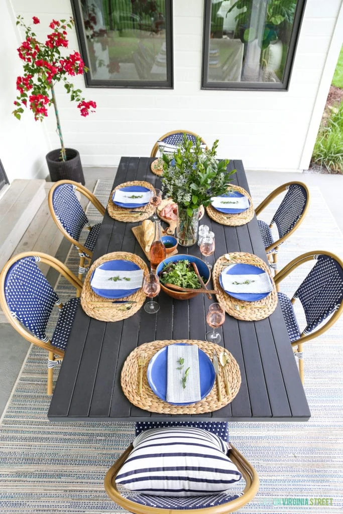 Outdoor dining tablescape with navy blue and white bistro chairs, wood table, striped pillow, seagrass chargers, blue melamine plates and wood accents.