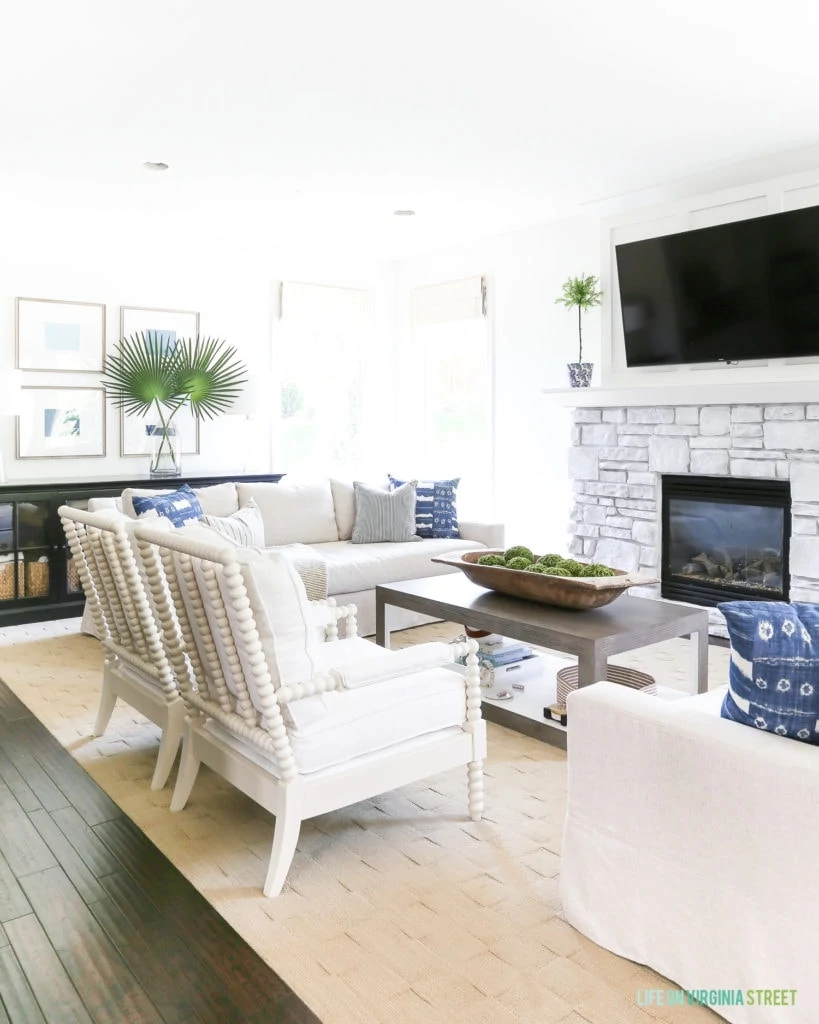 Coastal style living room with white, blue, green, and neutral accents. The space includes spindle chairs and linen slip-cover sofas. Blue and white shibori pillows are accented by striped pillows and ombre blue art. Wall color is Benjamin Moore Simply White.