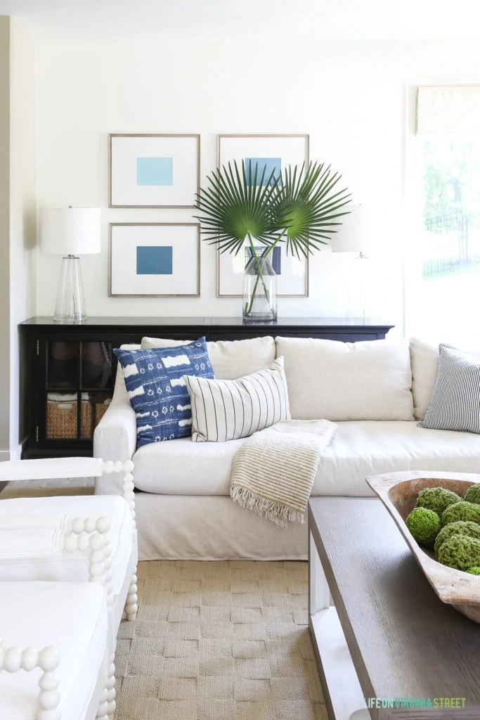 Ombre blue art in driftwood toned frames hang over a linen sofa with blue and white pillows. Accented by palmetto palm leaves.