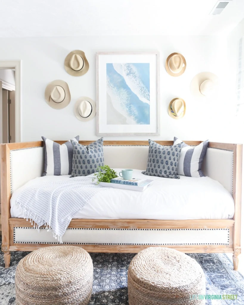 Beach artwork hanging over a wood and linen daybed. This white room is decorated with straw hats on the wall, round jute poufs and a dark blue patterned rug.