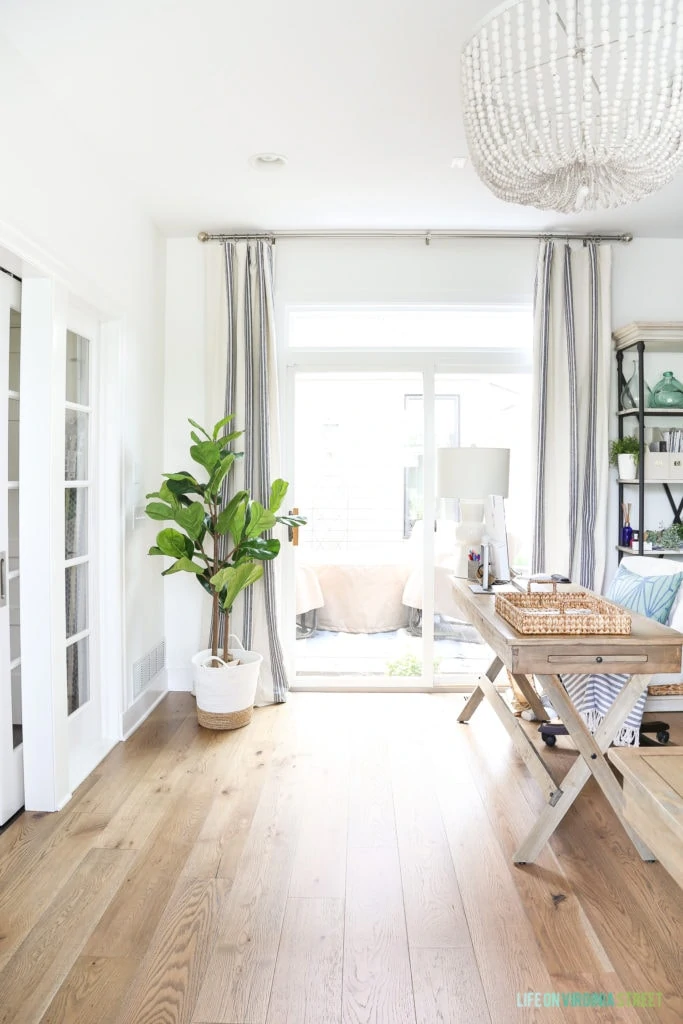 A home office space with white walls and trim, white oak hardwood floors, and white wood bead chandelier and a faux fiddle leaf fig tree. So many great tips for styling faux fiddle leaf fig trees and stems!