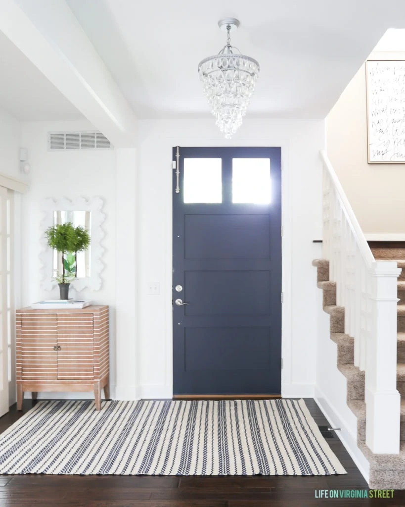 Summer entry with Benjamin Moore Simply White walls and Haley Navy door. The space includes a crystal chandelier, striped wood cabinet, green topiary, and striped rug.