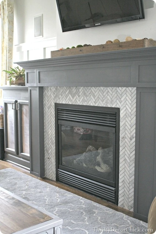 Grey and white fireplace with wooden box on top of mantel and TV on wall.