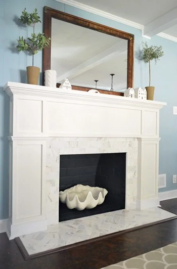White fireplace with large mirror above the mantel and topiaries on top.